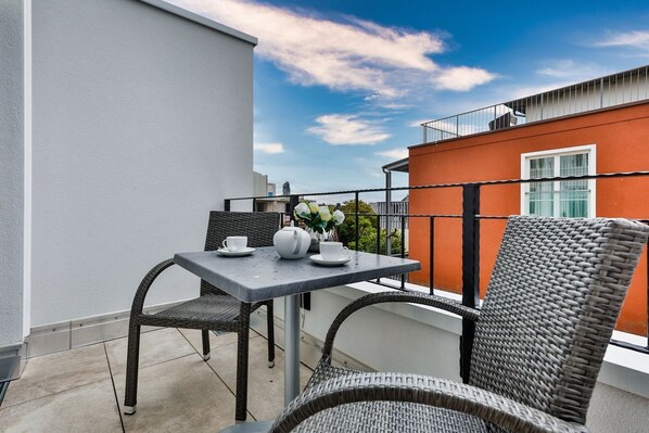 A balcony with a table and chairs, a potted plant, and a view of a red building.