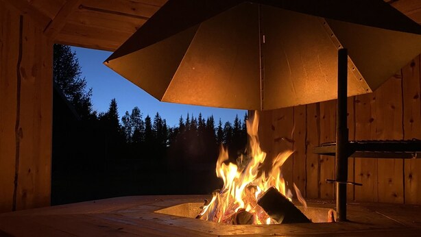 Una chimenea de leña con un paraguas grande y abierto encima, rodeada de paredes de madera y vistas a árboles al anochecer.