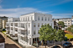 A multi-story white building with balconies, a flag, and a small garden area. - Logierhaus Jonna Whg. 15 - Logierhaus Jonna (Sylt)