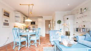 A spacious living room with a blue sofa, a dining table with chairs, and a kitchen in the background. - House Sönshörn, Germany (Sylt)