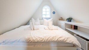 A bedroom with a sloped ceiling, a large window, a white bed with pillows, a nightstand, and a shelf with books and a vase. - House Sönshörn, Germany (Sylt)