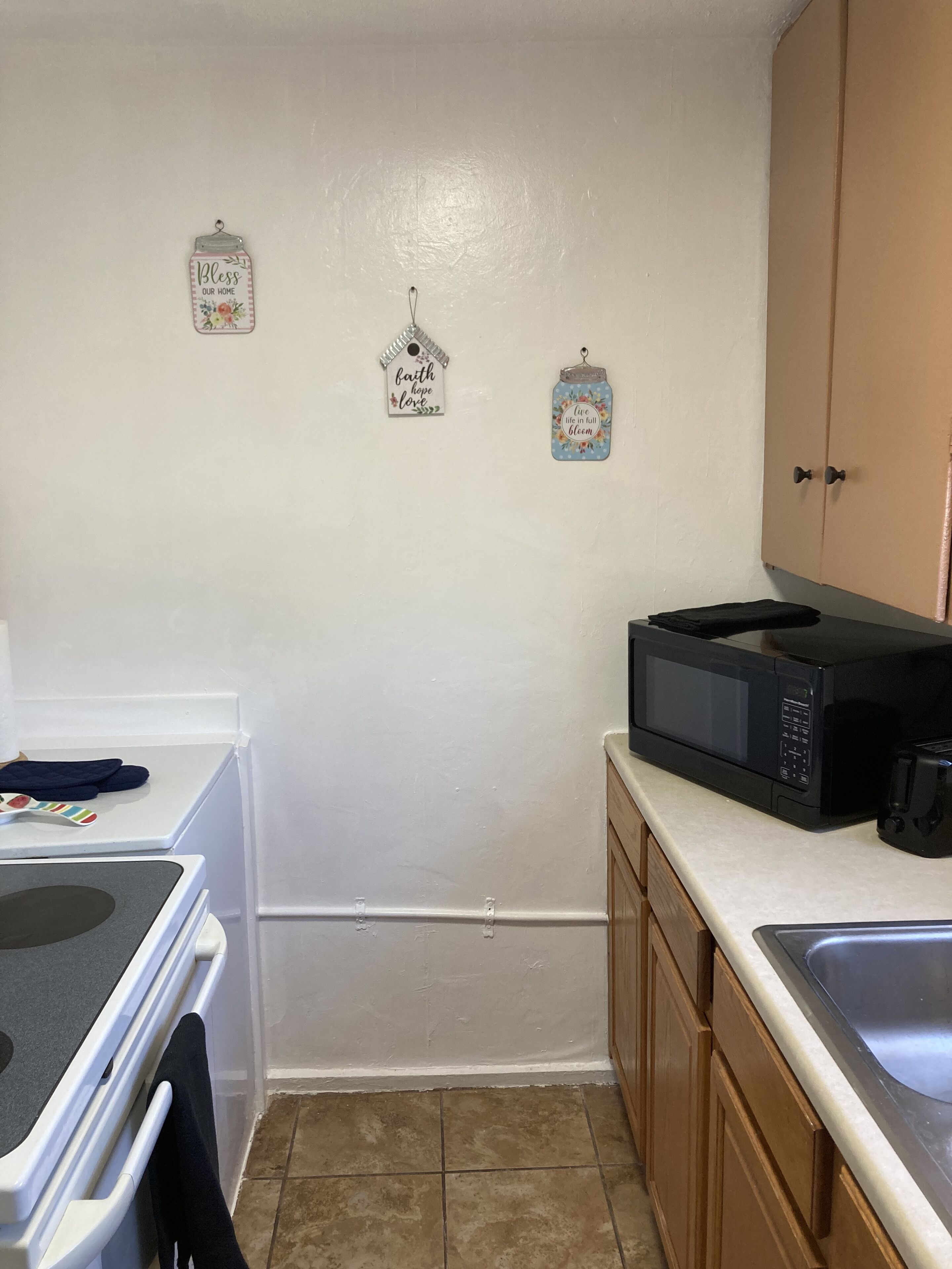 Une petite cuisine aux murs blancs, équipée d’un four à micro-ondes, d’une cuisinière et d’un évier. On y trouve également deux éléments décoratifs suspendus au mur et un meuble de rangement.