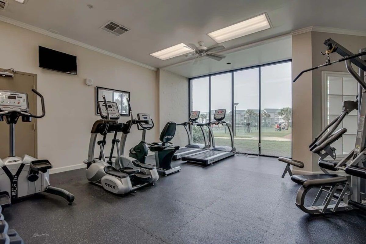 A gym with treadmills, elliptical machines, and a stationary bike, large windows, and a ceiling fan.