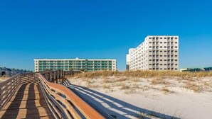 Een houten looppad leidt naar een hotel aan het strand, met een modern ontwerp, meerdere balkons en een helder uitzicht op de oceaan.