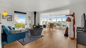 A spacious living room with a blue sofa, a grey armchair, and a dining area with wicker chairs. A large window offers a view of greenery and a building. - Cozy home in Hurup Thy with WiFi (Hurup Thy)