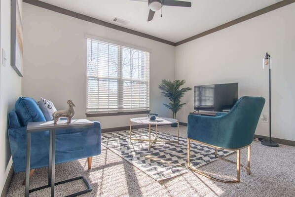 Un salon avec un canapé bleu, un fauteuil turquoise, une télévision, une petite table et un tapis à motifs.