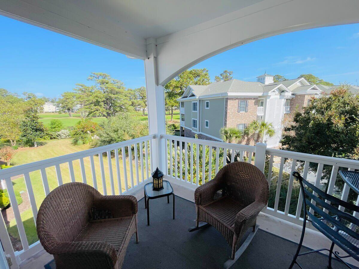 A covered balcony with wicker chairs and a small table, overlooking a well-maintained lawn and residential buildings.