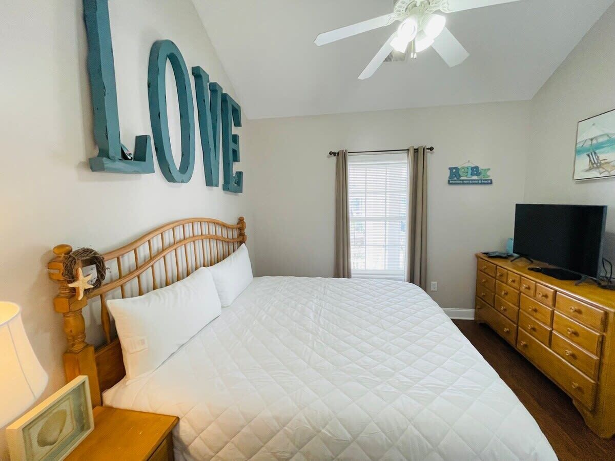 A bedroom with a large bed, a wooden headboard, a television on a wooden dresser, and a ceiling fan.