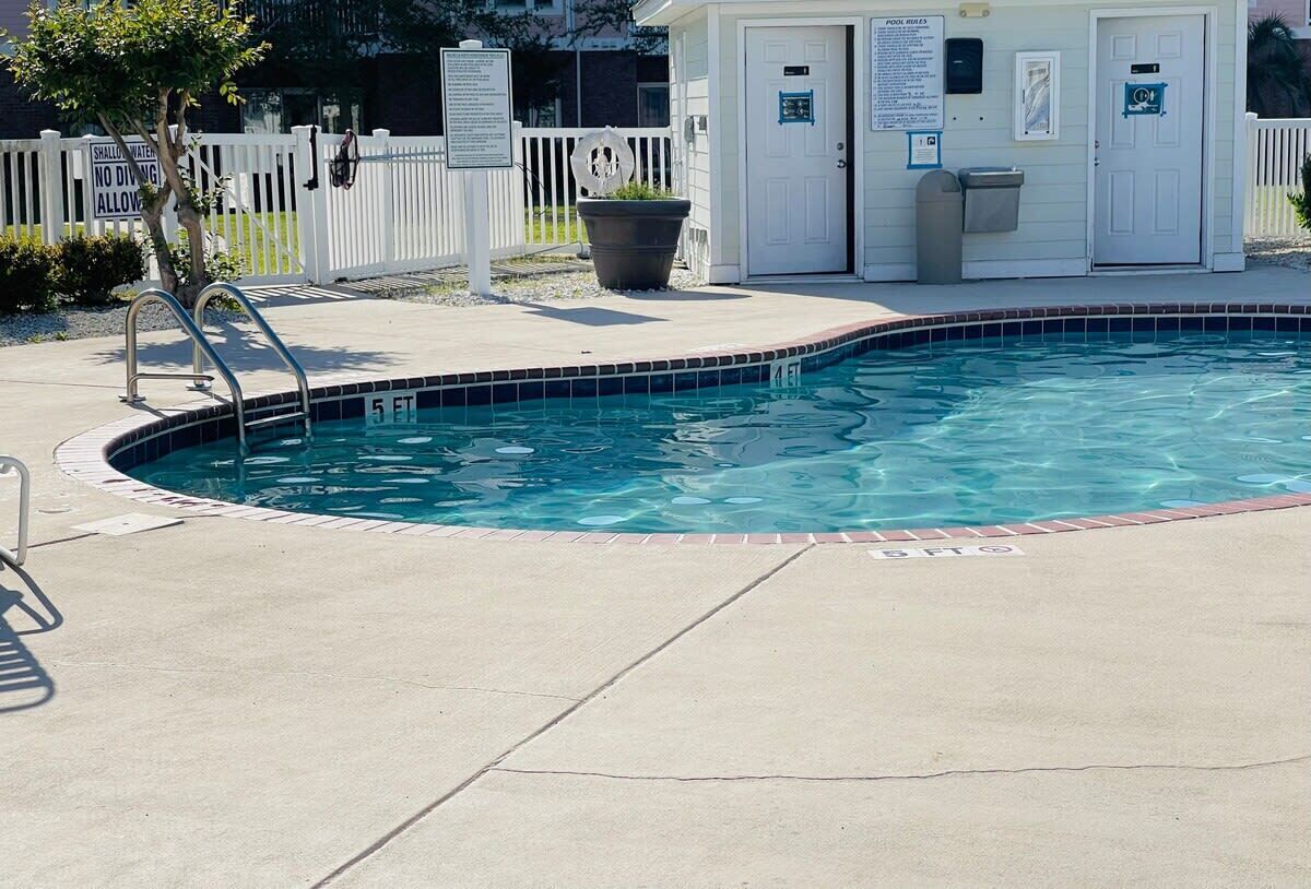 Une piscine entourée d’une clôture blanche, un petit bâtiment avec une porte et une poubelle.