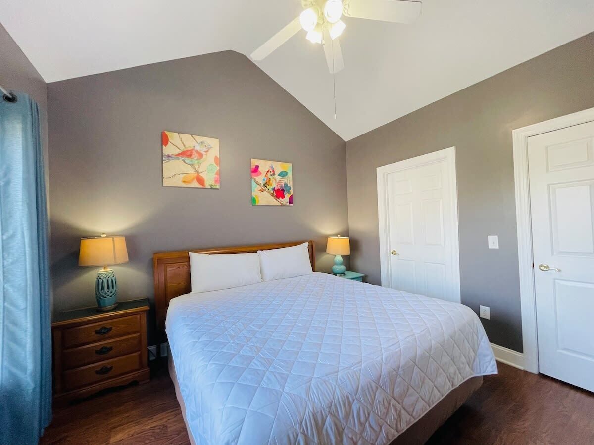 Une chambre à coucher comprenant un lit, deux tables de chevet, un ventilateur de plafond et deux tableaux accrochés au mur.