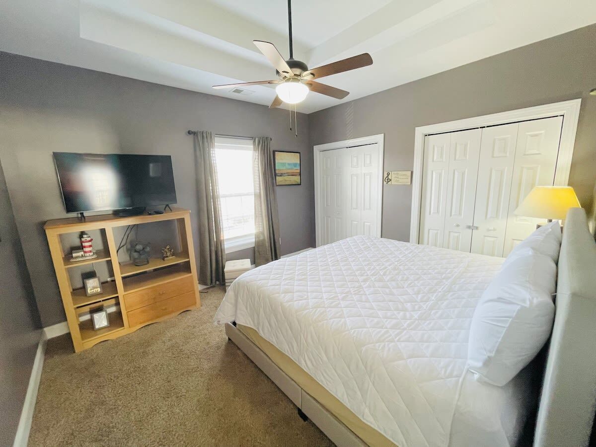 A bedroom with a bed, a TV on a wooden stand, a ceiling fan, and a door.