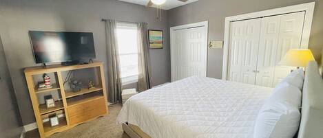 A bedroom with a bed, a TV on a wooden stand, a ceiling fan, and a door.