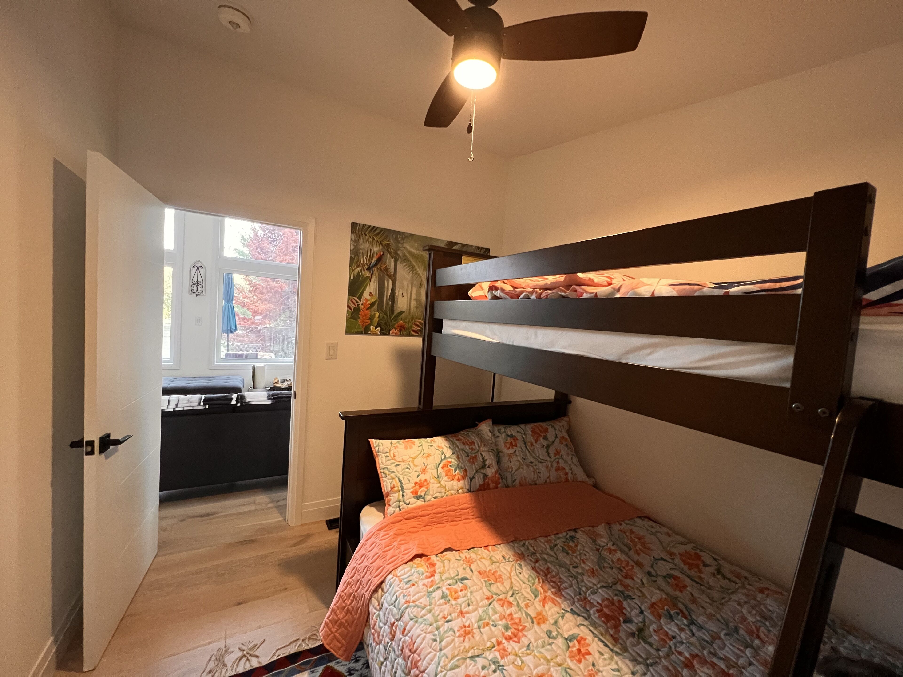 Een slaapkamer met een stapelbed, een plafondventilator, een raam met uitzicht op bomen en een deur die naar een andere kamer leidt.