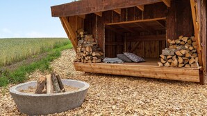A rustic wooden cabin with a porch, a fire pit, and a stack of firewood.