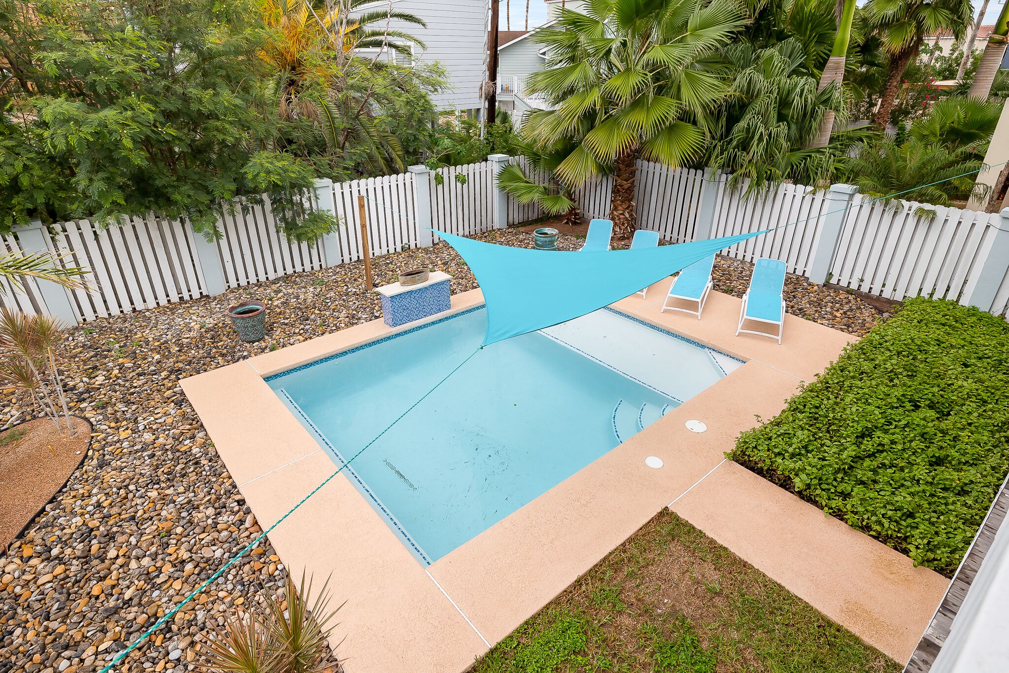 A rectangular swimming pool with a blue shade sail, surrounded by a white picket fence, pebbles, and small plants.