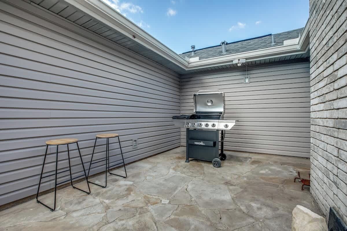 Een terras met een barbecue en krukken, stenen vloer, en een overdekt gedeelte met een dakraam.