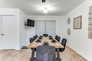 A dining area with a wooden table set for six, surrounded by chairs, with a television mounted on the wall and a door to the left.