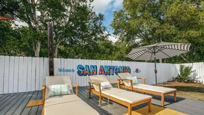 Une terrasse avec des chaises longues et un parasol à rayures, une clôture blanche sur laquelle on peut lire les mots « SAN ANTONIO » et « Bienvenue », et des arbres en arrière-plan.