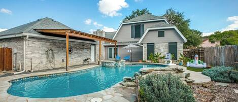 A backyard with a swimming pool, stone patio, and a wooden pergola.