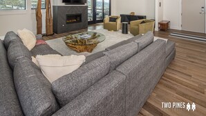 A modern living room with a large gray sectional sofa, a glass coffee table, and a fireplace. There are two windows offering a view of a snowy mountain landscape. - Steps to the Gondola | Ski-In/Ski-Out Luxury Penthouse (Big Sky)