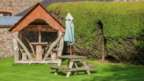 A rustic wooden structure with a picnic table and umbrella, surrounded by a well-maintained lawn and stone buildings in the background.