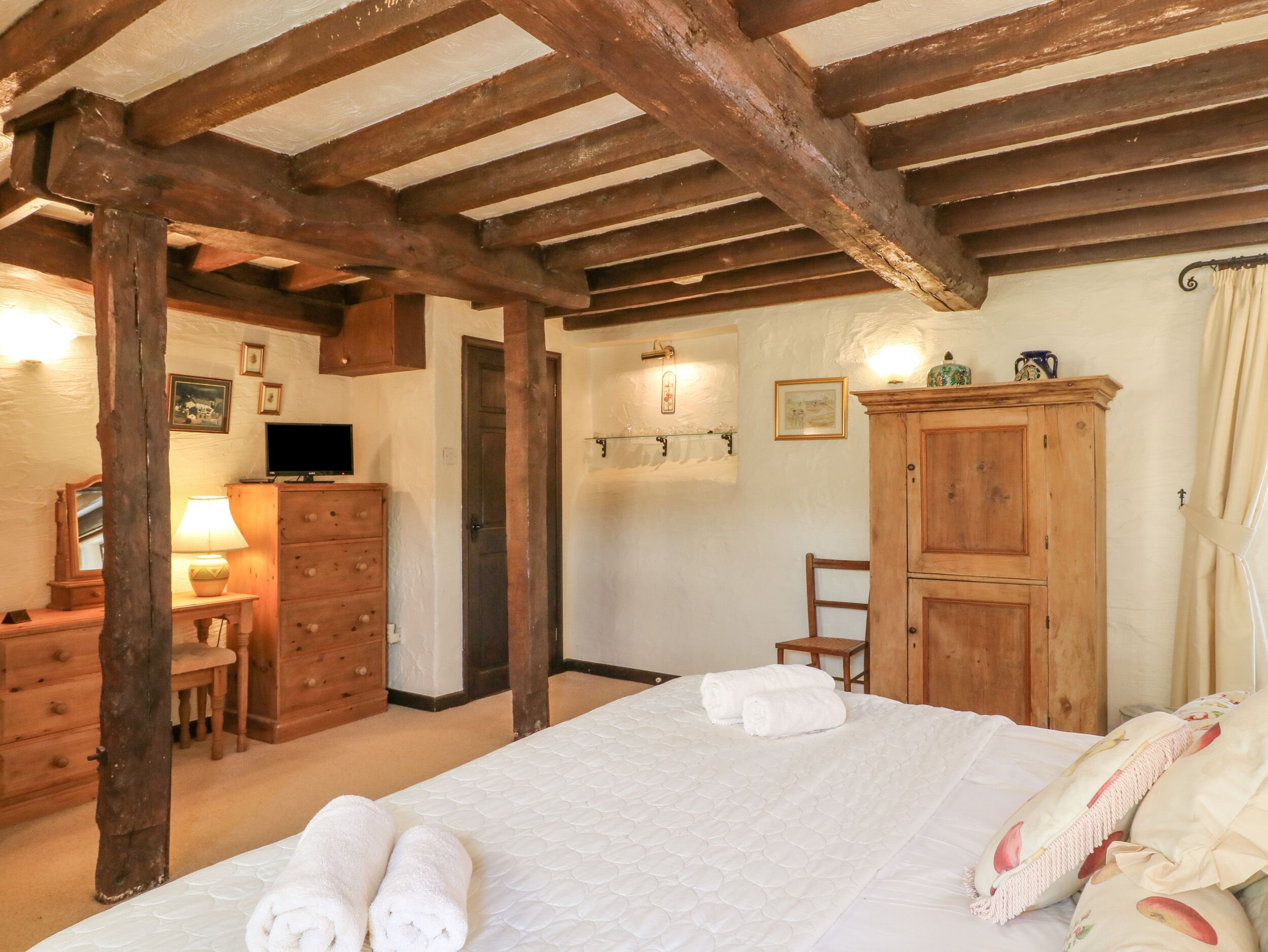 A bedroom with a wooden ceiling, a bed with white bedding, a wooden wardrobe, a desk with a lamp, and a television.