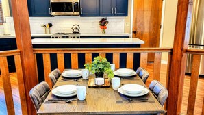 A dining area with a wooden table set for six, surrounded by chairs. There is a kitchen in the background with blue cabinets and a microwave.