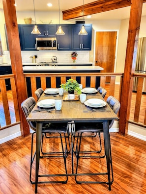 Une salle à manger avec une table en bois dressée pour six personnes, entourée de chaises. On aperçoit au fond une cuisine avec des placards bleus et un four à micro-ondes.