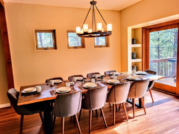 Un comedor con una mesa de madera dispuesta para seis personas, rodeada de sillas, una araña y vistas a árboles a través de amplios ventanales.