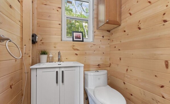 Une petite salle de bain aux murs lambrissés, avec un lavabo blanc, des toilettes et une fenêtre située au-dessus du lavabo.