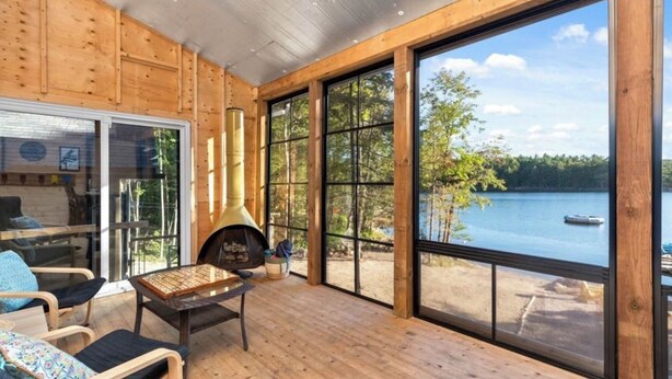 A cozy cabin with a large window overlooking a lake, a fireplace, and wooden walls.