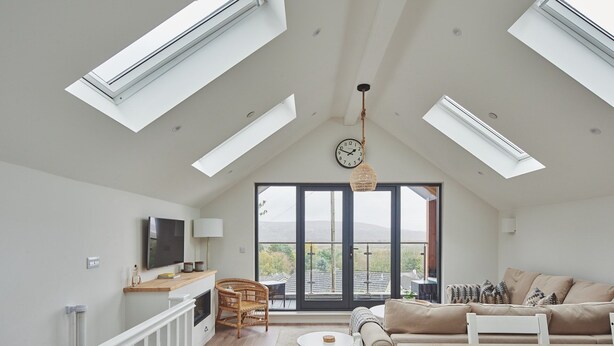 A spacious living room with a high ceiling, skylights, a large window, a sofa, a TV, and a dining area. - Ty Florence - Luxury Cottages (Swansea)