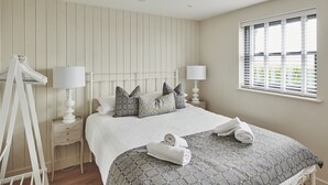 A bedroom with a bed, two bedside tables, a window with blinds, and a hanging rack. - Ty Florence - Luxury Cottages (Swansea)