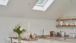 A modern kitchen with a skylight, white cabinetry, a dining area with white chairs, and a coffee station. - Ty Florence - Luxury Cottages (Swansea)