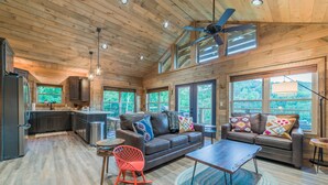 A spacious living area with a brown leather sofa, a red rocking chair, and a coffee table. There are large windows providing natural light and a view of the outdoors. - Fun and Funner! Family cabin with mountain view and pinball! (Sevierville)