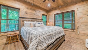 A wooden cabin bedroom with a bed, two windows with blinds, a ceiling fan, and a small wooden stool. - Fun and Funner! Family cabin with mountain view and pinball! (Sevierville)