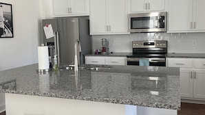 A modern kitchen with white cabinets, a stainless steel refrigerator, a microwave, and a built-in oven. There are three woven stools at the island. - Oxford Townhouse Minutes from campus! (Oxford)