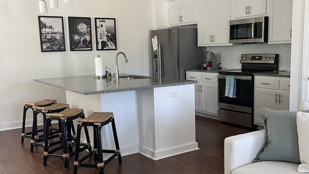 A modern kitchen with white cabinets, stainless steel appliances, and a central island. There are three bar stools and a dining table with a checkered tablecloth. - Oxford Townhouse Minutes from campus! (Oxford)