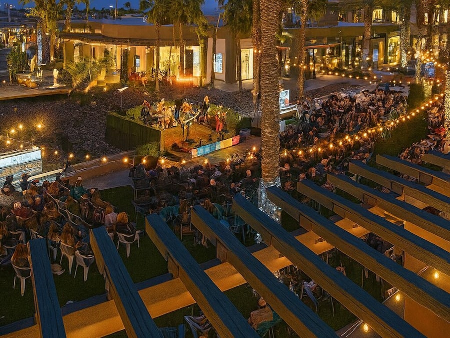 An outdoor seating area with rows of chairs and tables, palm trees, and a stage for performances.