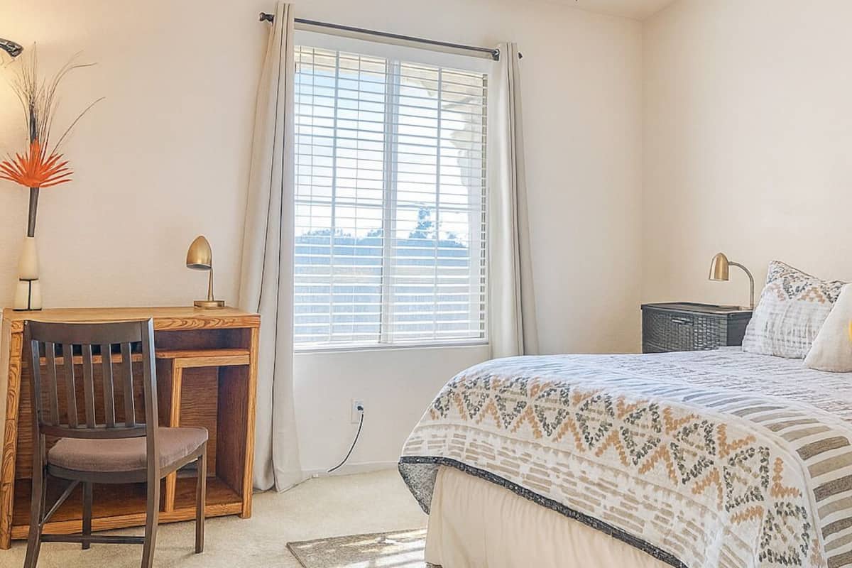 A bedroom with a bed, a desk with a chair, a ceiling fan, and a window with blinds.