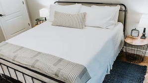 A bed with a metal frame, white bedding, striped pillows, a small bedside table with a lamp, and a rug. - GrueneGusthaus#1| Private Condo +Golf Cart Rentals (New Braunfels)