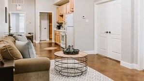 A living room with a sofa, a coffee table, and a patterned rug. There is a kitchen area with wooden cabinets and a refrigerator. - GrueneGusthaus#1| Private Condo +Golf Cart Rentals (New Braunfels)