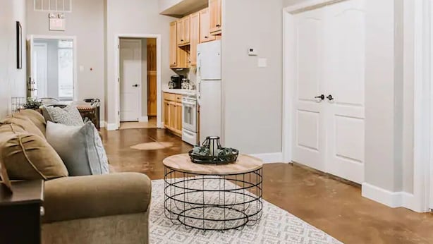 A living room with a sofa, a coffee table, and a patterned rug. There is a kitchen area with wooden cabinets and a refrigerator. - GrueneGusthaus#1| Private Condo +Golf Cart Rentals (New Braunfels)