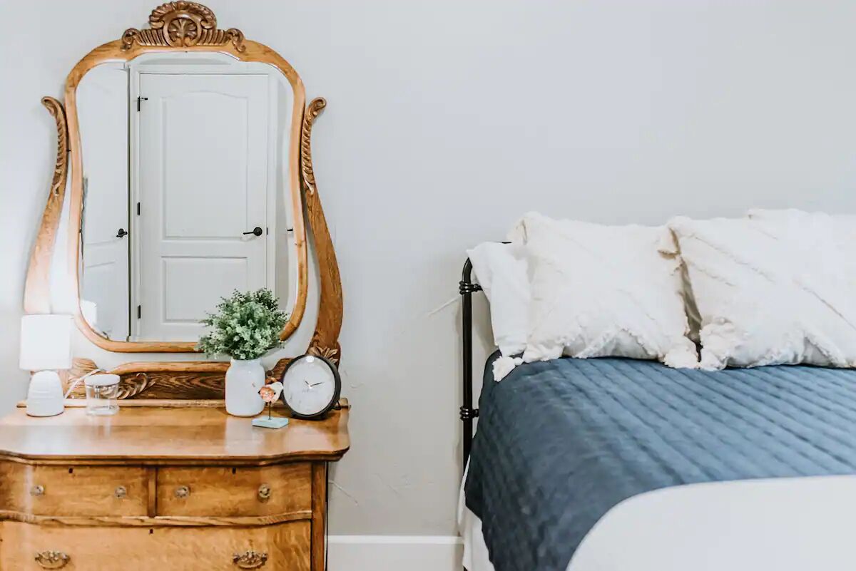 Un dormitorio con un aparador de madera, un espejo, un reloj y una cama con almohadas blancas y una colcha azul.