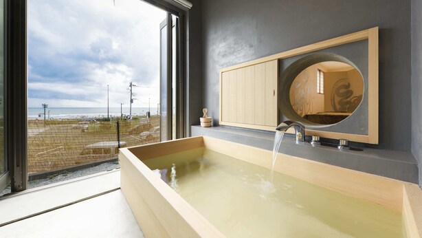 A modern bathroom with a large bathtub, a wooden vanity, and a large window offering a view of the beach. - Yuigahama Chiyo (Kamakura)