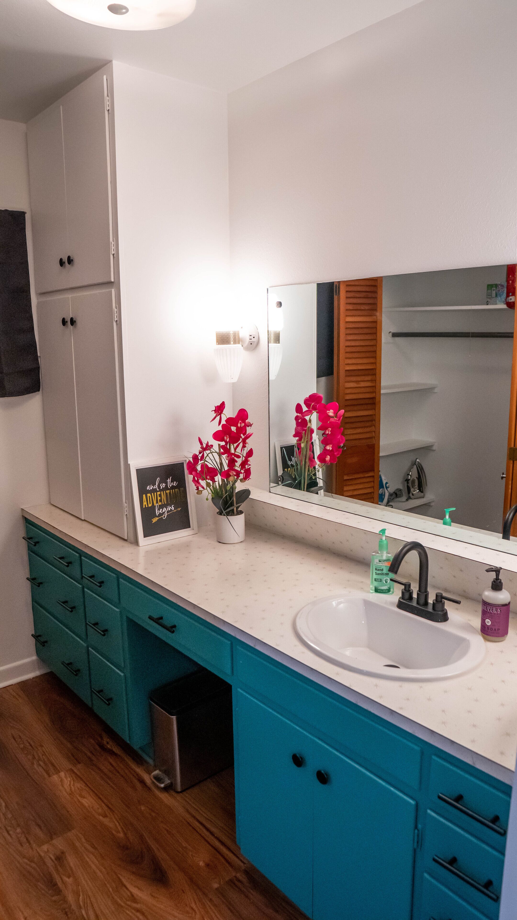Un bagno con lavandino bianco, mobili color verde acqua, uno specchio e un'orchidea in vaso.