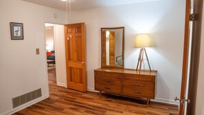 Quarto com cômoda de madeira, espelho e luminária. Uma porta dá acesso a outro cômodo.