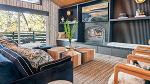 A cozy living room with a fireplace, wooden furniture, and a large window overlooking a deck. - Ski View Chalet - Super Chic & Modern, Hot Tub, Shuffleboard, Views (Big Bear Lake)