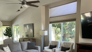 A living room with a grey sofa, a glass coffee table, and two white armchairs. There is a large window with blinds, a ceiling fan, and a flat-screen TV mounted on the wall. - Warm and inviting home centrally located. (La Quinta)
