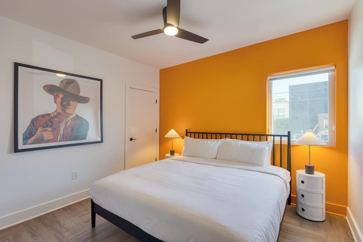 A bedroom with a bed, a ceiling fan, a nightstand with a lamp, a framed picture on the wall, and a window with a view of buildings.